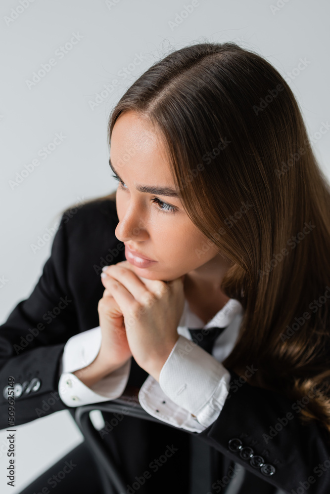high angle view of young and brunette woman in suit leaning on wooden chair back isolated on grey. high angle view of young and brunette woman in suit leaning on wooden chair back isolated on grey.
