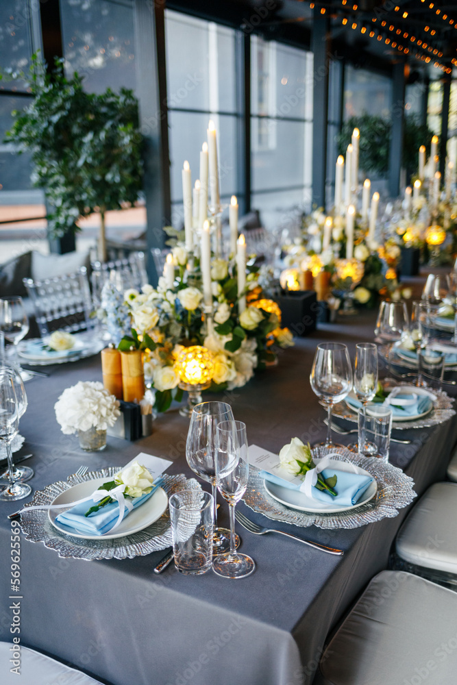 Table setting decorated with a gray tablecloth, silver plates, blue napkins and compositions of white, blue flowers and candles. Concept of wedding decorations 