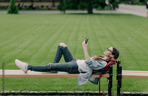 Young beautiful woman student lying on a banch in a campus listening to music or audio lectures.