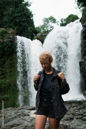 A young blonde girl with a backpack travels to a large waterfall.