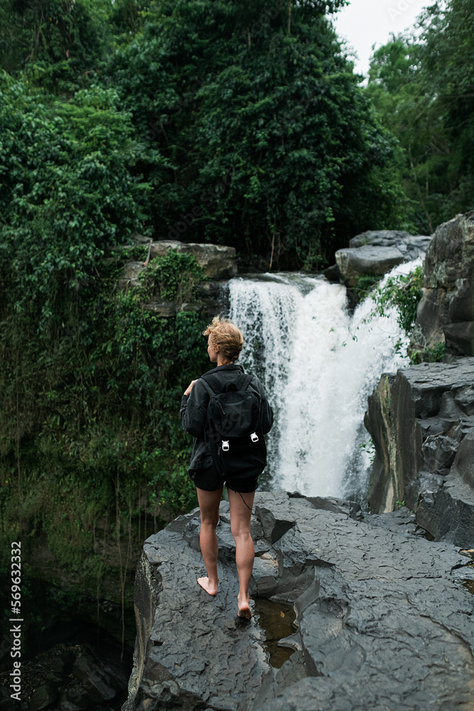 A young blonde girl with a backpack travels to a large waterfall, rear view.