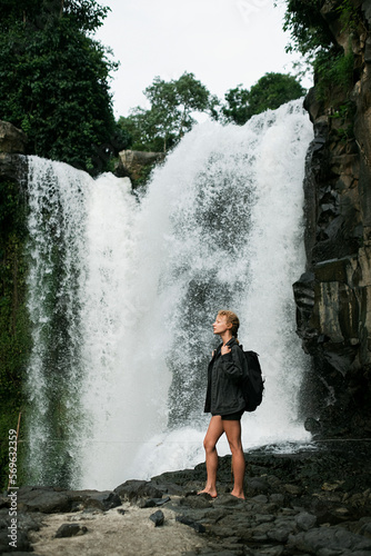 A young blonde girl with a backpack travels to a large waterfall.