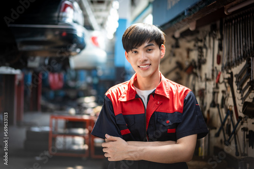 Selective focus, front view of a young handsome Asian male mechanic in red and blue uniform, standing with arms crossed and smiling at the camera with blurred lifted cars in garage in the background.