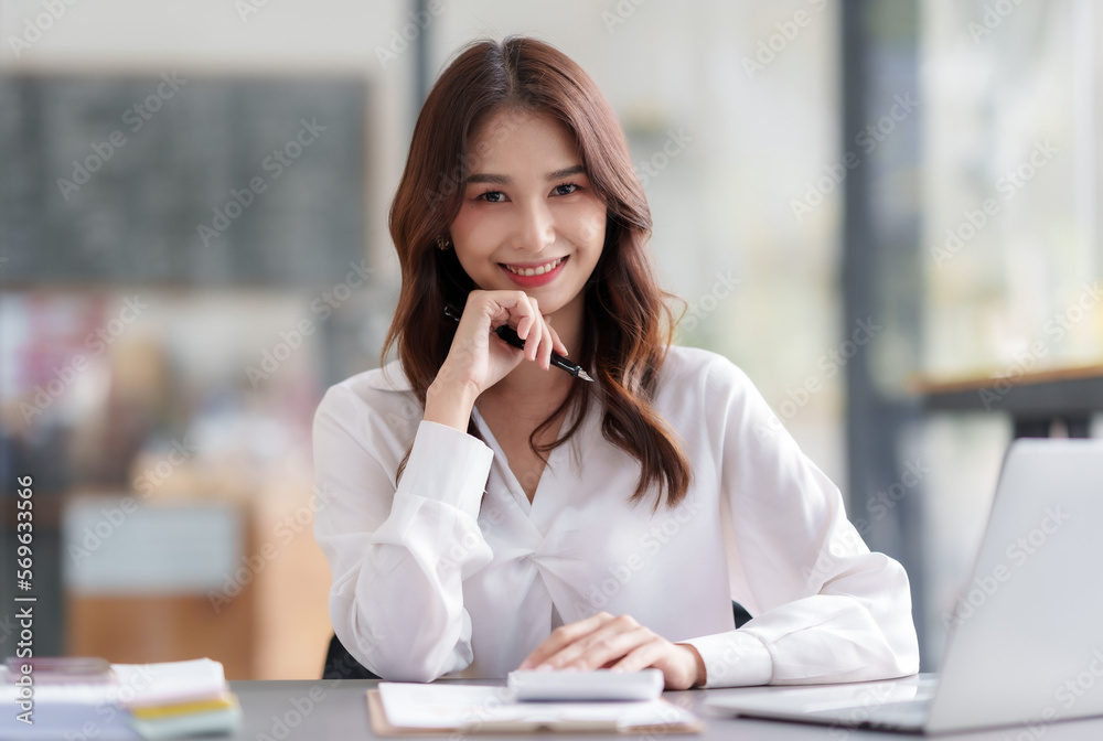 Pretty Asian businesswoman sitting smiling and happy working on laptop computer and document in office. Looking at camera. Pretty Asian businesswoman sitting smiling and happy working on laptop computer and document in office. Looking at camera.