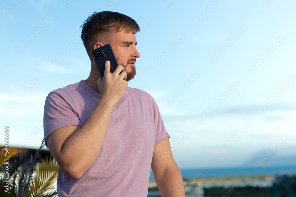 Happy young handsome man is talking on cell mobile phone, calling on smartphone, having conversation with friend, smiling outdoors at summer sunny day
