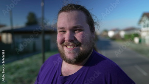 One joyful young chubby person stands outdoors smiling at camera. Overweight male person closeup face