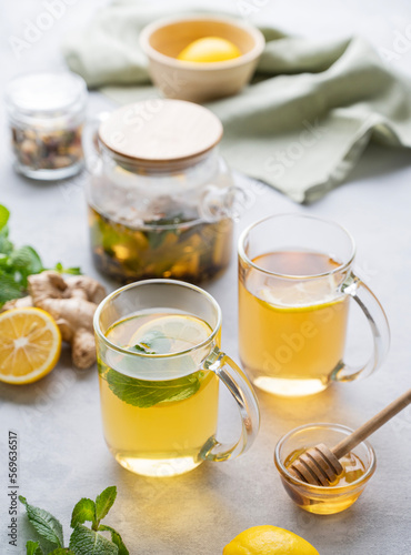 Two glasses of tea with fresh lemon, honey, mint and ginger on a light background with teapot and morning light close up.