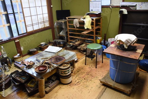 A traditional urushi craftsman's workshop in Japan with tools, wooden trays, bowls, and materials spread on a well-used table. Shelves, containers, and shoji screens complete the rustic workspace.