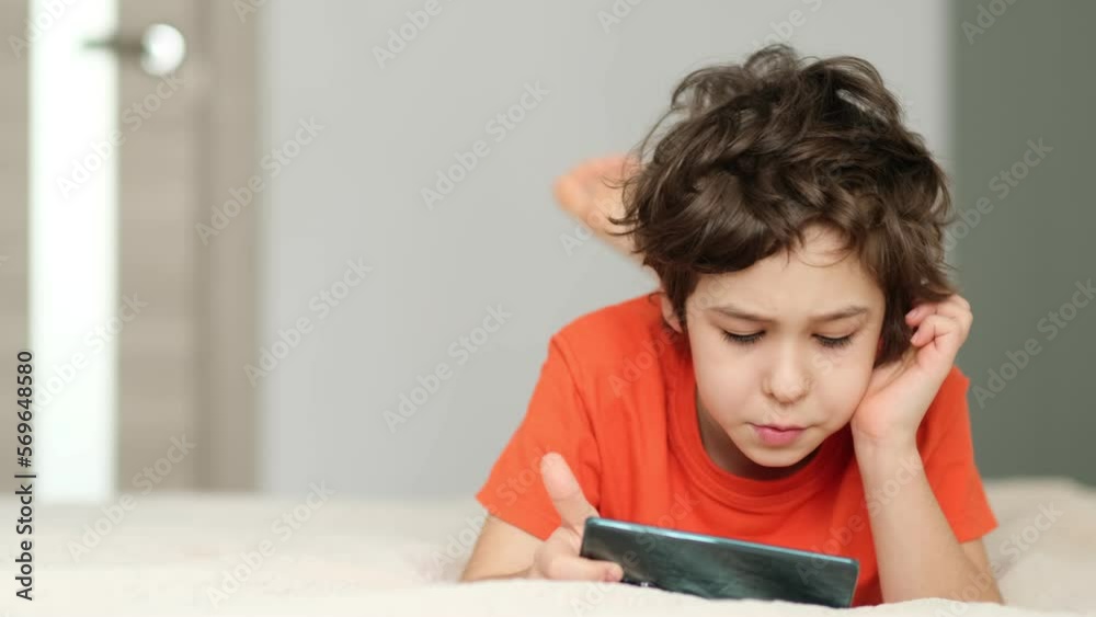 primary school boy plays on the phone. child on a bed under a blanket uses gadgets. preteen boy looks at the screen primary school boy plays on the phone. child on a bed under a blanket uses gadgets. preteen boy looks at the screen