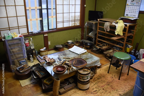 A traditional urushi craftsman's workshop in Japan with tools, wooden trays, bowls, and materials spread on a well-used table. Shelves, containers, and shoji screens complete the rustic workspace.