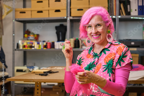 Seamstress with pink hair and colorfull clothes hand sewing a piece of fabric, smiling and looking at camera, in a sewing workshop.