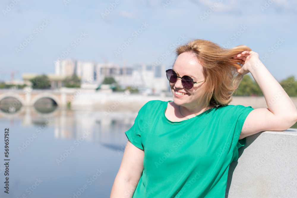 Portrait of a girl in sunglasses, a green blouse standing on the riverbank holding her hair with her hand