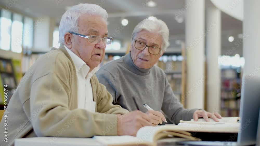 Focused senior men writing abstract in notebook in library. Side view of gray-haired friends studying together, talking, devoting time to hobby and education. Friendship, retirement concept Focused senior men writing abstract in notebook in library. Side view of gray-haired friends studying together, talking, devoting time to hobby and education. Friendship, retirement concept