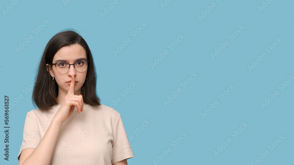 Portrait of a serious student girl with glasses, covering her mouth with her finger on a blue background. Portrait of a serious student girl with glasses, covering her mouth with her finger on a blue background.