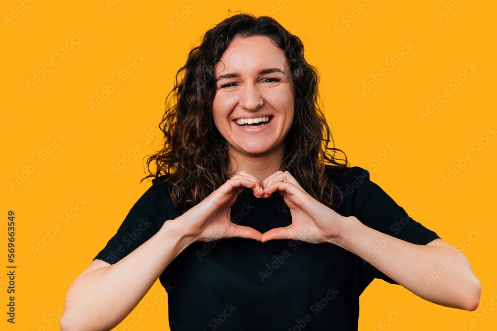 Close up portrait of a laughing woman showing heart gesture over her chest over yellow background.