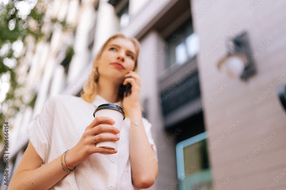 Low-angle view of young pretty woman in casual clothes talking on smartphone while standing on city street on summer day. Elegant female teenager with coffee to go calling taxi by mobile phone.