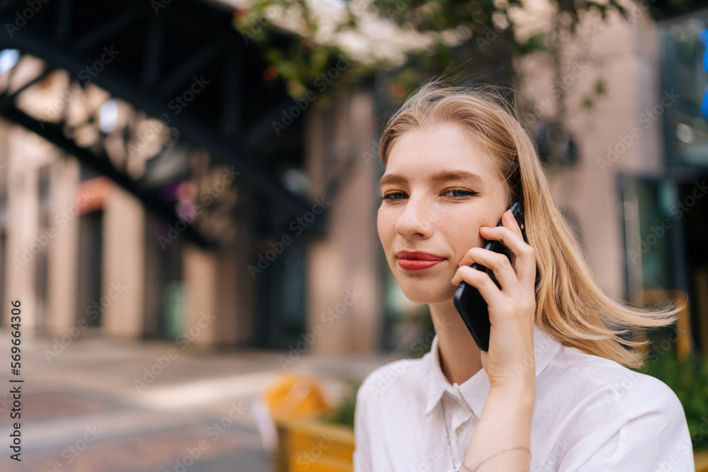 Close-up portrait of happy blonde young woman talking phone and smiling looking at camera standing on city street. Front view of gorgeous positive female using mobile phone outdoors. Close-up portrait of happy blonde young woman talking phone and smiling looking at camera standing on city street. Front view of gorgeous positive female using mobile phone outdoors.