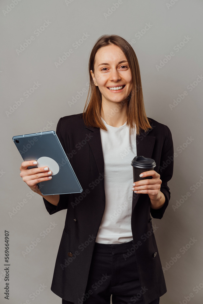 Confident woman wearing jacket holding a tablet and a to go cup of coffee over grey background.