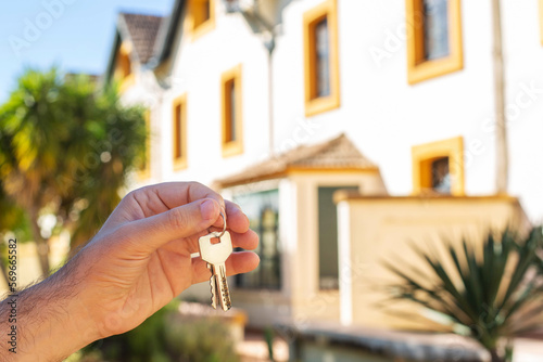 A man's hand holds the keys to the apartment against the background of the house and palm trees. The topic of buying expensive real estate in countries with a warm climate.
