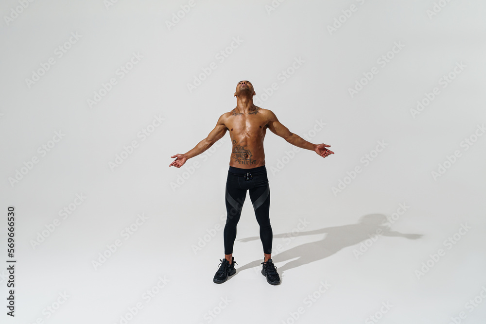 Attractive sporty man with muscled naked body on white studio background looks up with open arms