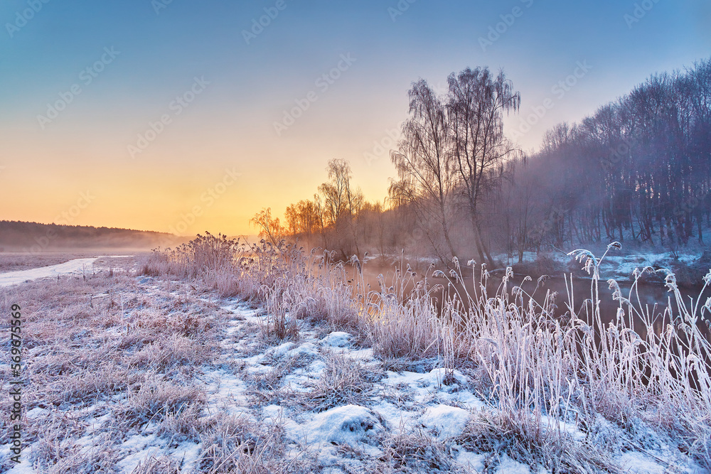 Dawn on winter riverbank. Frost on grass, cane. Snowy rural road. January fog. Forest river sunrise. Cold Weather landscape, reflection in water.