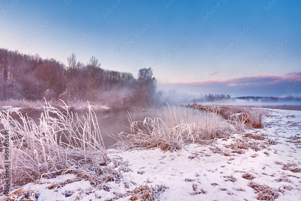 Dawn on winter riverbank. Frost on grass, cane. Snowy rural road. January fog. Forest river sunrise. Cold Weather landscape, reflection in water
