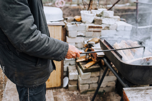 A man in the open air, in nature, holds a metal poker in his hands and corrects burning firewood in the barbecue. Photography, food preparation.