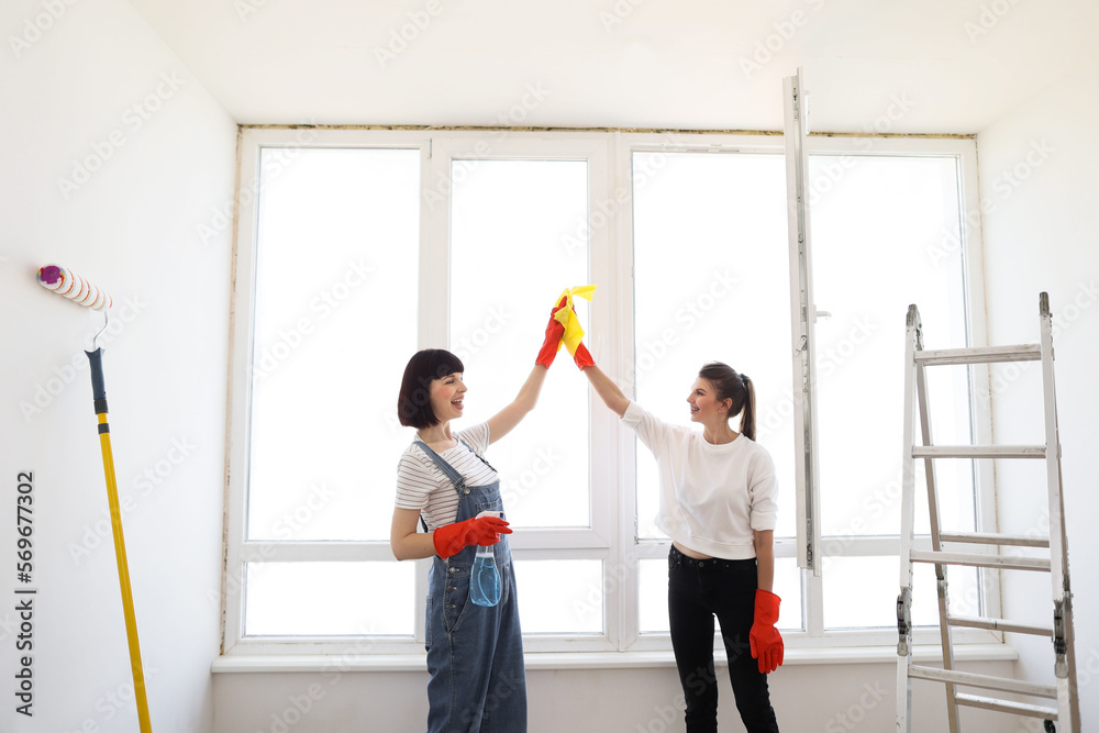 Fototapeta premium Beautiful young women in rubber gloves with detergent and rag washing windows together giving high five. Attractive caucasian girls making house cleaning after repair.