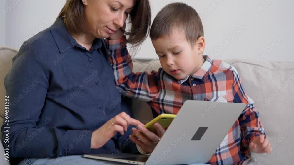 Mother and Her Little Son Using Phone and Laptop Sitting on Couch. Woman Shows Boy Video or Mobile Games on Smartphone Mother and Her Little Son Using Phone and Laptop Sitting on Couch. Woman Shows Boy Video or Mobile Games on Smartphone