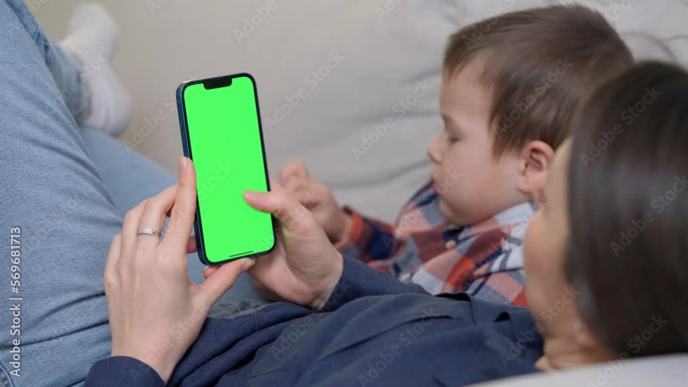 Mother Lying on Sofa with Kid and Using Smartphone with Green Screen, Chroma Key. Boy Plays Mobile Games On Phone. Scrolling Through Social Media or Online Shop Mother Lying on Sofa with Kid and Using Smartphone with Green Screen, Chroma Key. Boy Plays Mobile Games On Phone. Scrolling Through Social Media or Online Shop