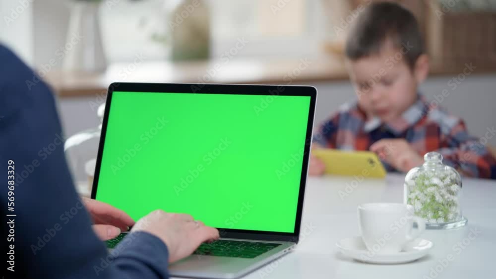 Woman Uses Laptop with Green Mock-up Screen at Home. Background is Little Boy Plays Mobile Games, Watches Videos and Social Media On Phone Woman Uses Laptop with Green Mock-up Screen at Home. Background is Little Boy Plays Mobile Games, Watches Videos and Social Media On Phone