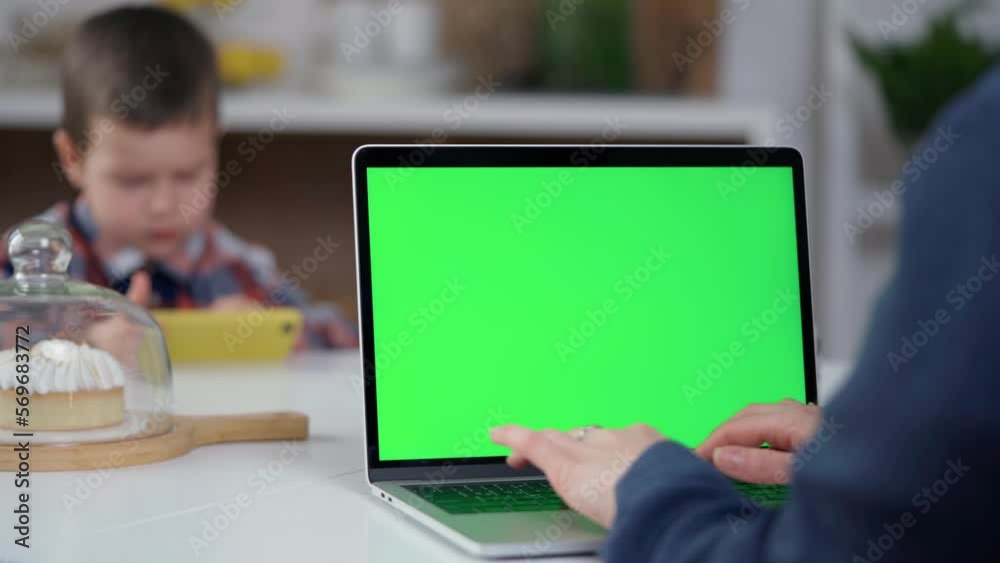 Woman Uses Laptop with Green Mock-up Screen at Home. Background is Little Boy Plays Mobile Games, Watches Videos and Social Media On Phone Woman Uses Laptop with Green Mock-up Screen at Home. Background is Little Boy Plays Mobile Games, Watches Videos and Social Media On Phone