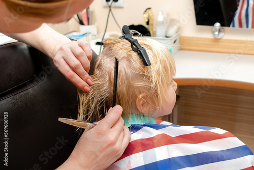 Little blond child boy getting haircut in beauty salon,barbershop. Hairdresser stylist cutting hair with professional tools-comb and scissors.Kids spa.Style and fashion,cutting hair for children