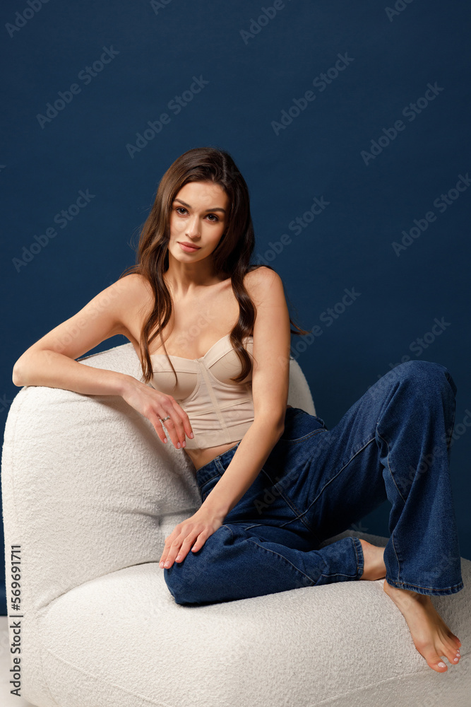 A beautiful stylish girl in blue jeans and a beige corset sits on a chair and looks into the frame. Blue background. A beautiful stylish girl in blue jeans and a beige corset sits on a chair and looks into the frame. Blue background.
