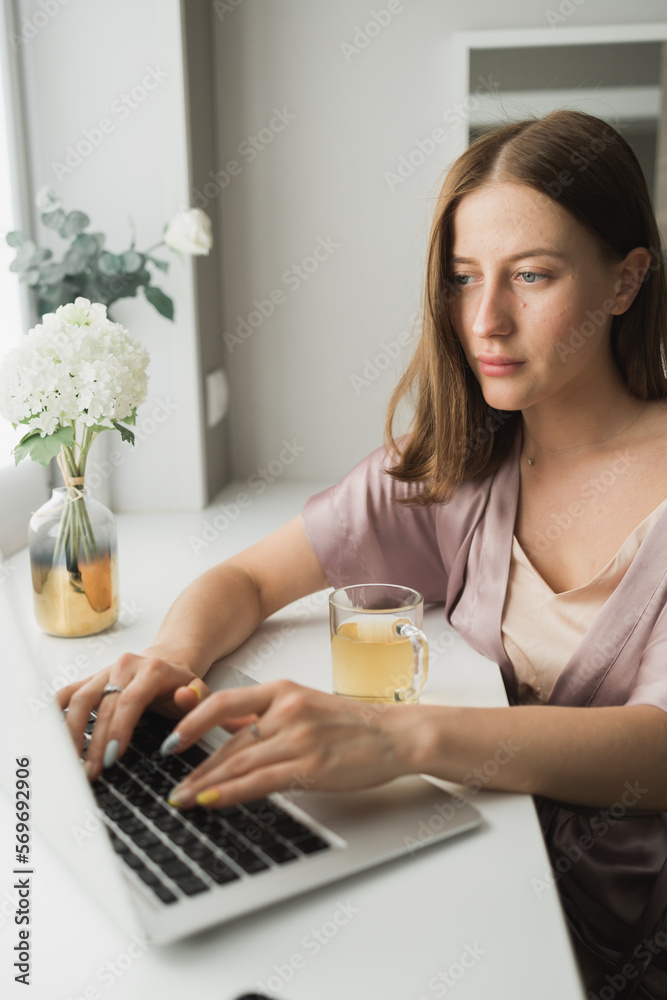 Young woman wears sleeping clothes working on laptop computer while sitting at living room and drinking tea - social network and working at home concept