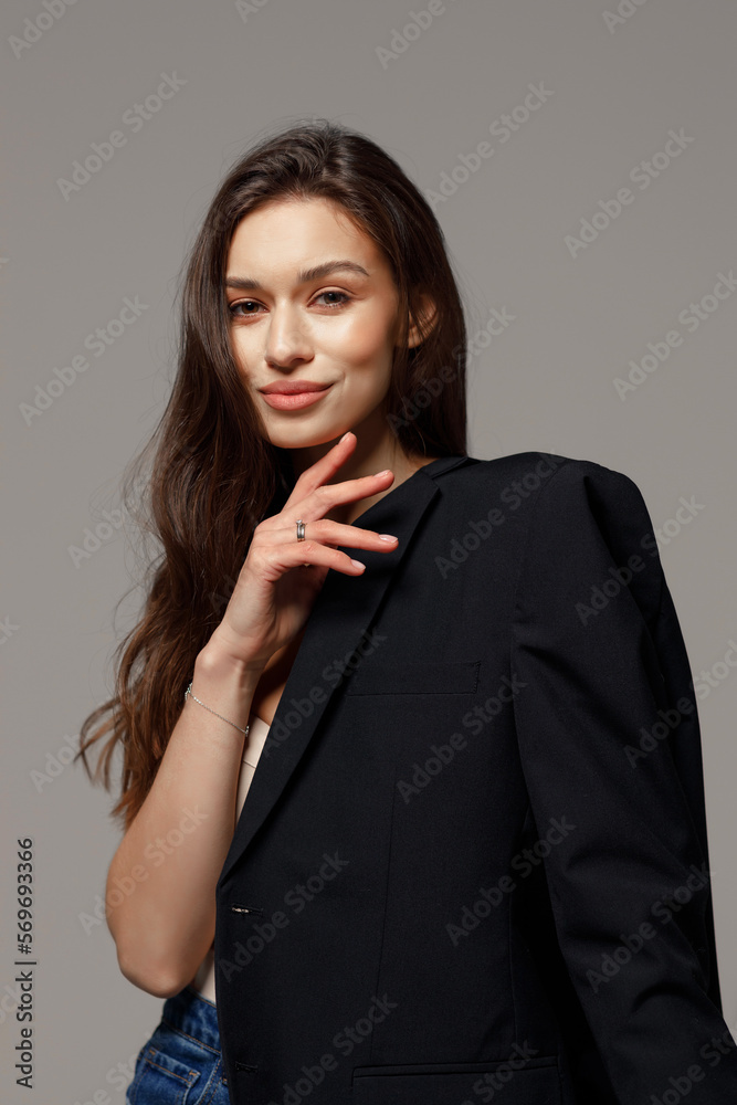 Beautiful stylish smiling girl in blue jeans and black jacket sitting on a chair on a gray background. The concept of lightness and femininity, fashion.