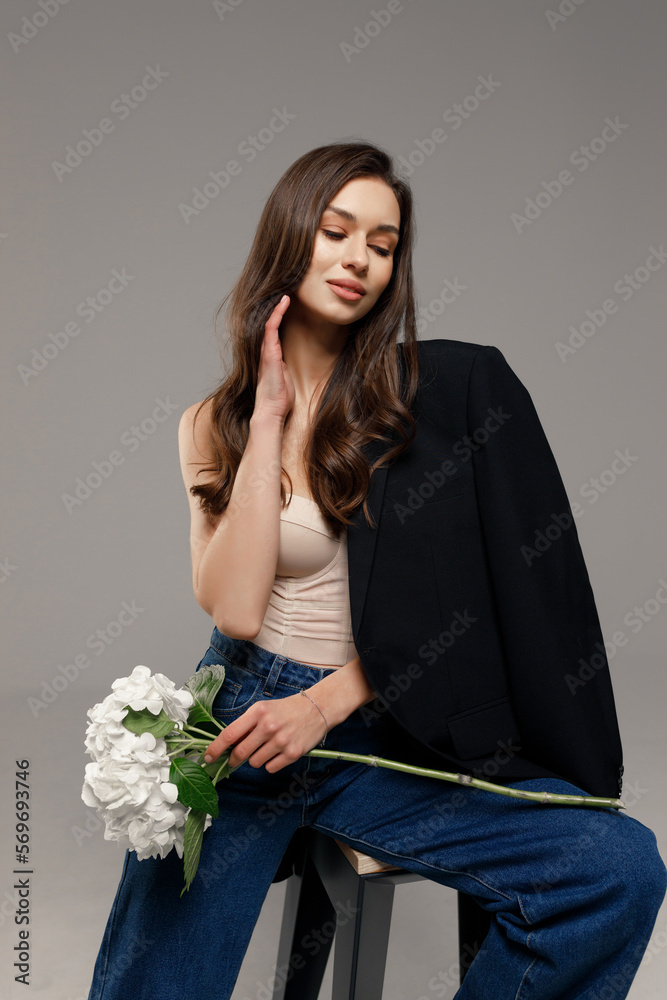 Φωτογραφία A beautiful stylish smiling girl in blue jeans and a black jacket sits on a chair on a gray background and holds white flowers in her hands