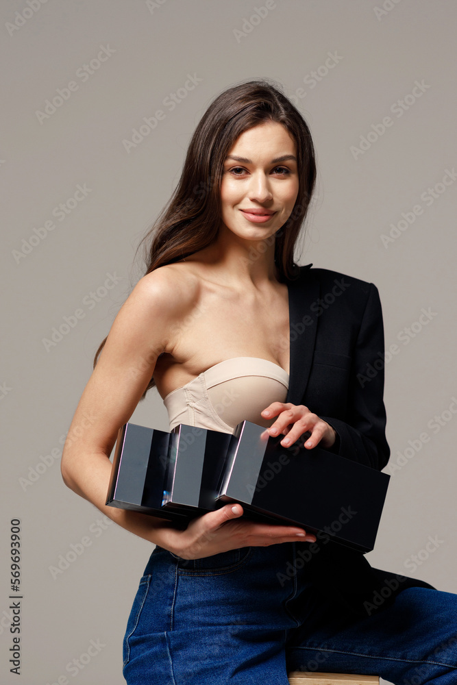 Φωτογραφία Beautiful stylish girl in blue jeans with bare shoulders and glowing skin, sitting on a chair on a gray background and holding cosmetic products in her hands