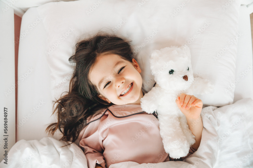 a little dark-haired girl is sleeping in bed, hugging a teddy bear under a white blanket.the child is in bed in the morning.