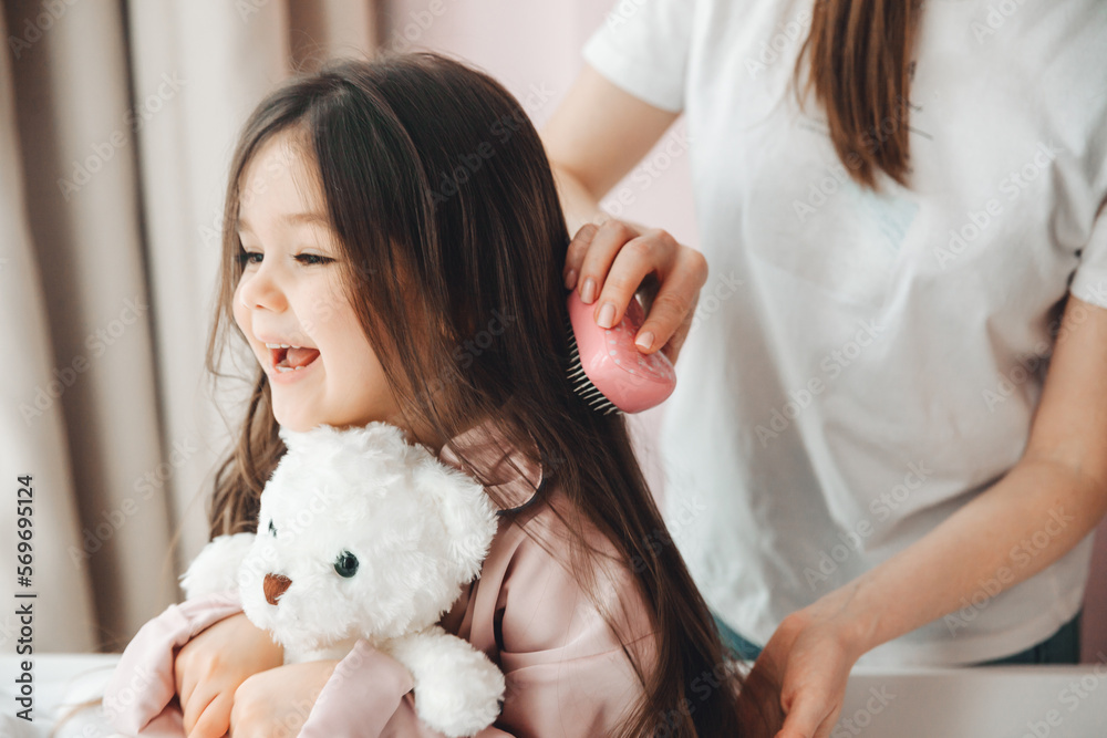 a little dark-haired girl looks into the camera and smiles while her mother combs her daughter's hair. hair care a little dark-haired girl looks into the camera and smiles while her mother combs her daughter's hair. hair care