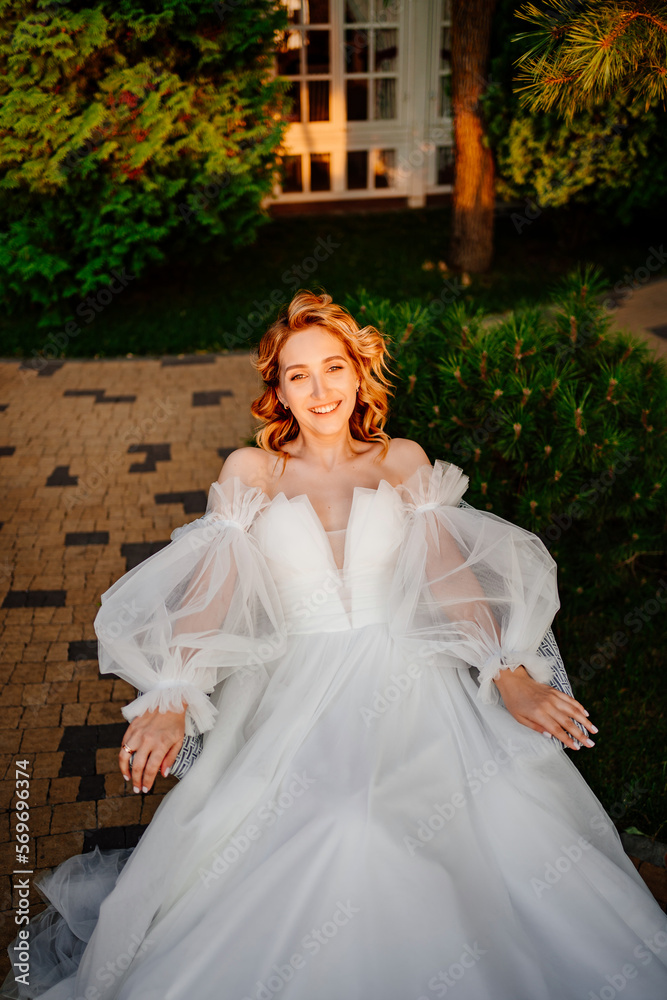 an attractive bride in a white delicate dress on a chair in the park at sunset. 