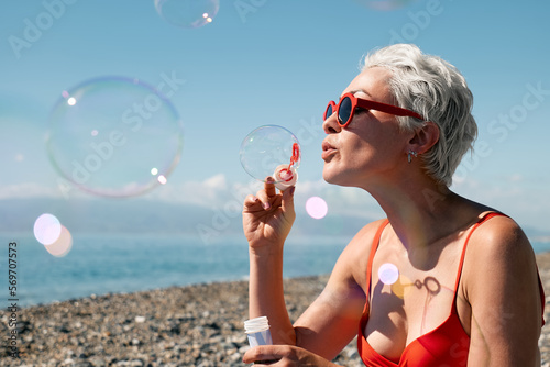 Blonde woman in sunglasses having fun and blowing soap bubbles on beach. Travel and beach lifestyle concept.