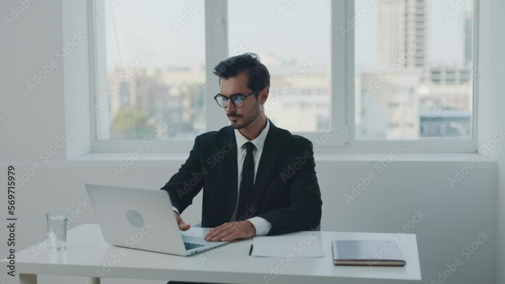Man business morning came to work in a suit with glasses with a smile sits behind a chair in the office opens a laptop and papers business documents, online work at the computer and papers documents. Man business morning came to work in a suit with glasses with a smile sits behind a chair in the office opens a laptop and papers business documents, online work at the computer and papers documents.