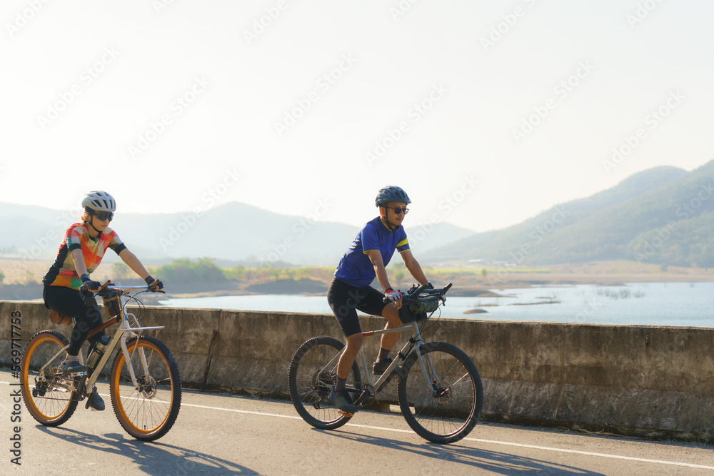 Obraz premium Asian cyclist couple riding together for exercise around the lake in the morning with beautiful mountain view in the background.