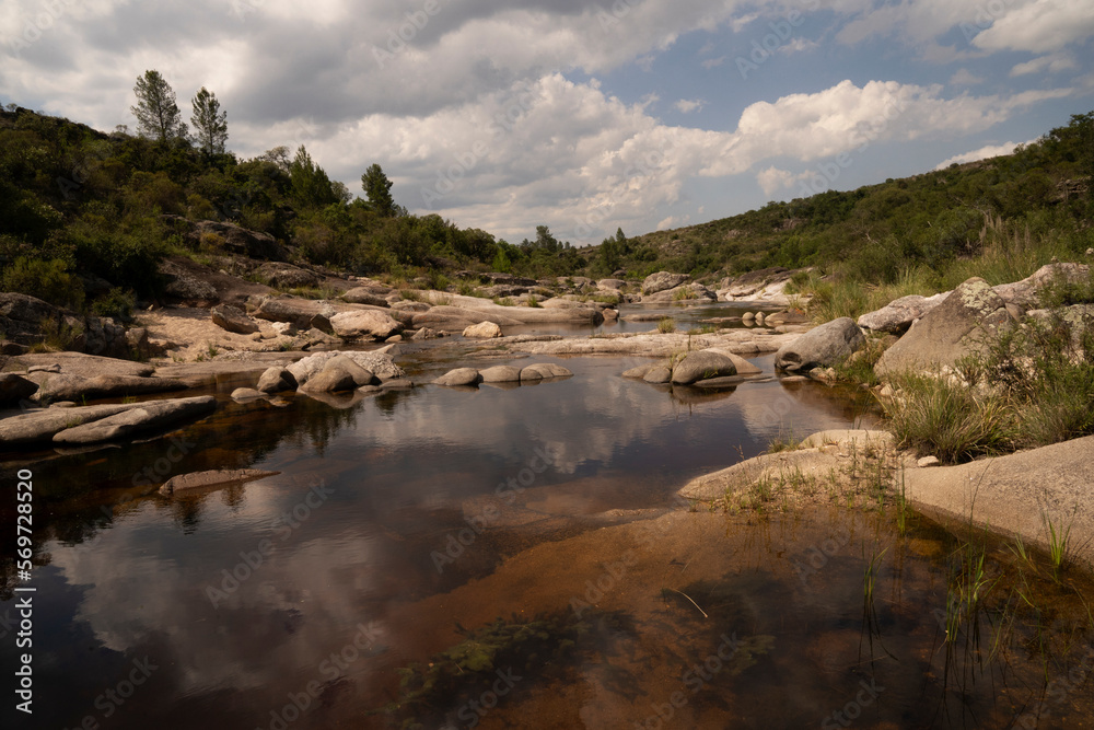 View of the calm river flowing across the rocky hills and forest. The blue sky with clouds reflection in the water surface. 