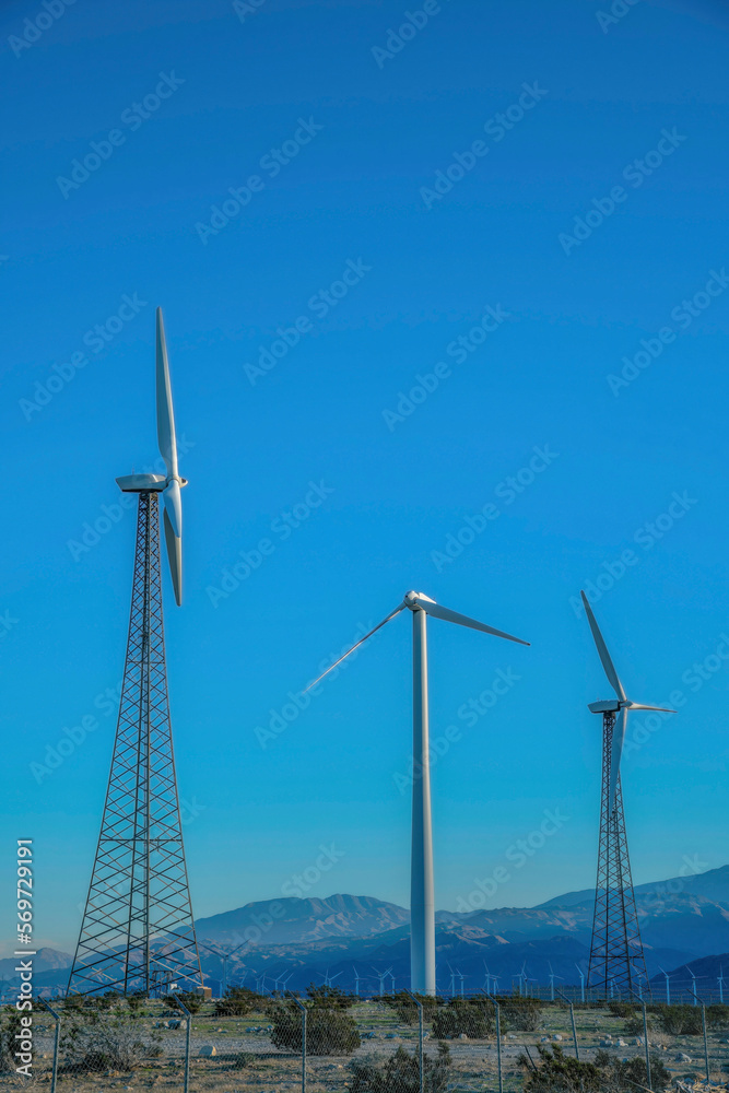Tubular steel wind turbine tower in the middle of two lattice wind turbine towers in California. There is a mesh wire fence at the front in a windmill farm on a desert.