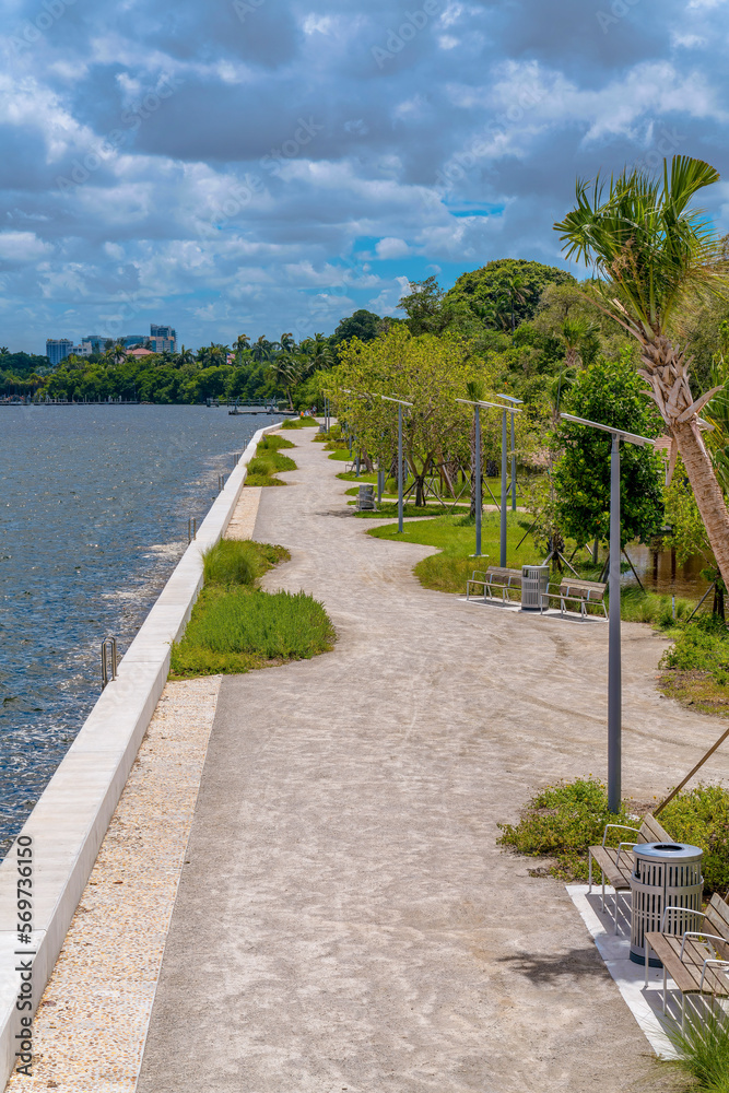 Alice Wainwright Park, Miami, Florida- concrete path near the water with green plants along the way. High angle view of a concrete path at shoreline with benches on the right against the cloudy sky.