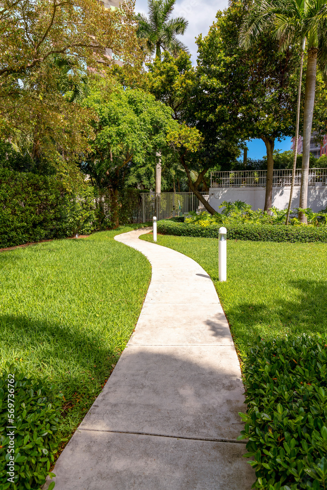 Concrete walkway at the entrance of a gated community in Miami, Florida. Narrow walkway in the middle of a green lawn with white lawn lights on the right heading to the white gate railings. Concrete walkway at the entrance of a gated community in Miami, Florida. Narrow walkway in the middle of a green lawn with white lawn lights on the right heading to the white gate railings.