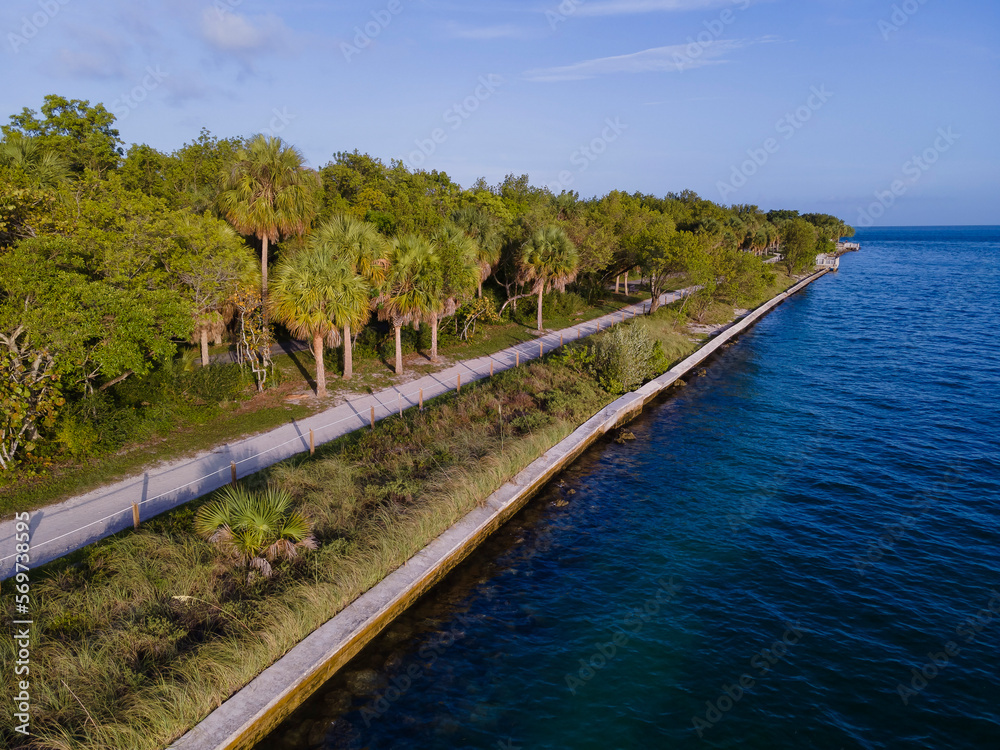 Road near the shore with concrete sea wall at Bill Baggs Cape Florida State Park in Miami, Florida. Aerial view of a road along the shore with trees and grasses near the blue ocean waters.