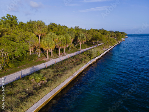 Road near the shore with concrete sea wall at Bill Baggs Cape Florida State Park in Miami, Florida. Aerial view of a road along the shore with trees and grasses near the blue ocean waters.